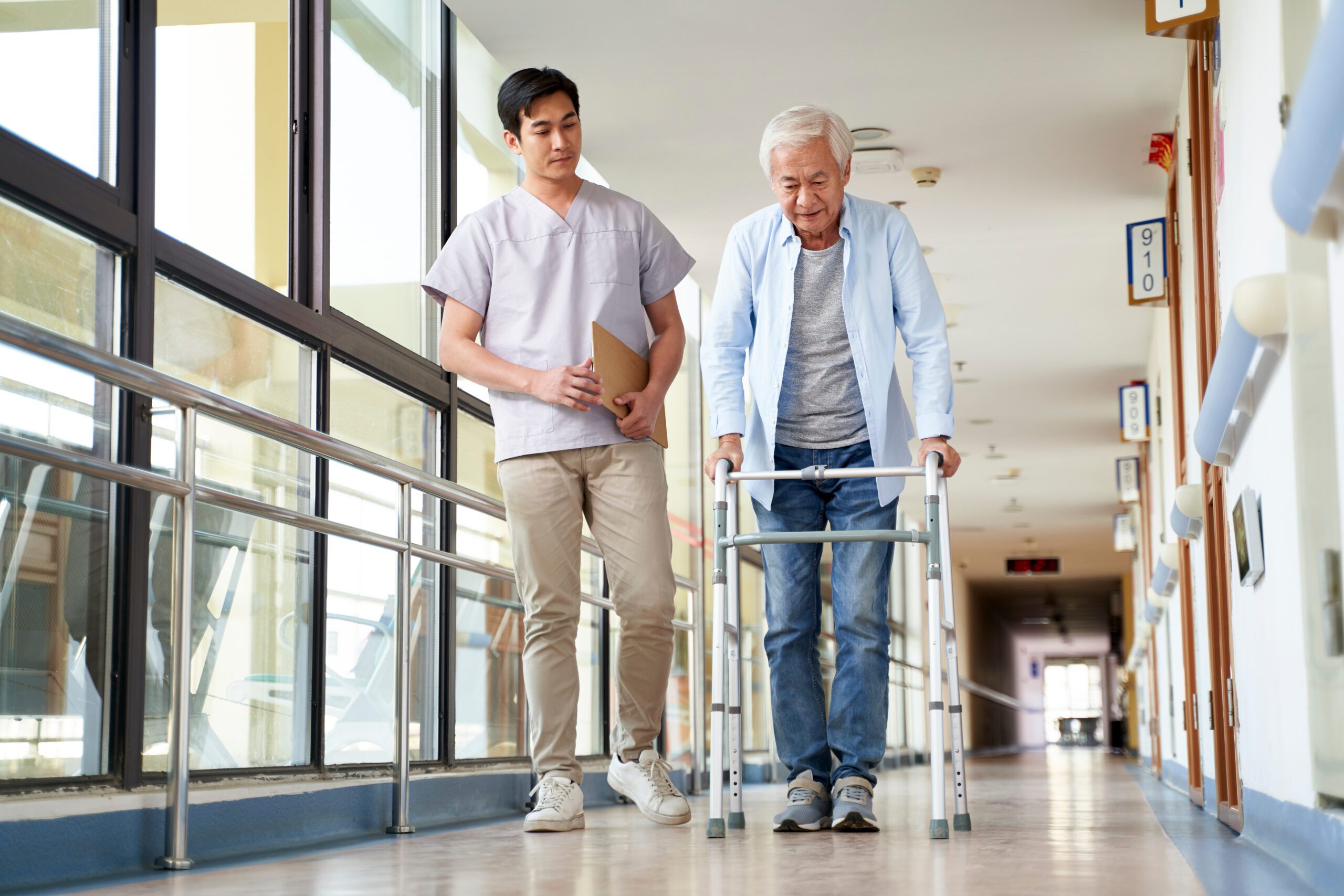 An elderly man with white hair using a walker, assisted by a young male caregiver holding a clipboard, walking down a bright hospital or care facility corridor.