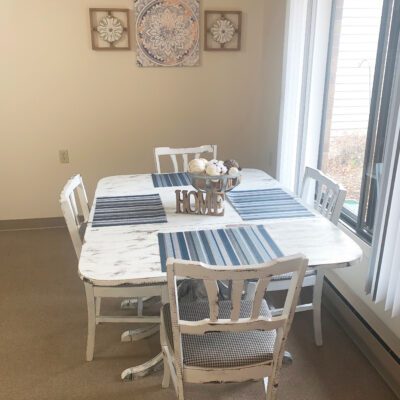 A dining room with a white table and six matching white chairs, placed near a large window with vertical blinds. The table is set with blue and white striped placemats and a decorative centerpiece that spells 
