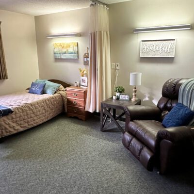 A cozy bedroom with a bed on the left, decorated with pillows and a quilt, and a wooden nightstand with a lamp, picture frame, and small plant. To the right, there is a dark leather armchair with a blue pillow, a side table with a white lamp and small plants, and wall decor including a 